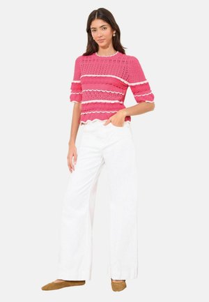 Frau trägt ein rosa kurzärmeliges Strickoberteil mit weißen wellenförmigen Streifen, weiße weit geschnittene Hosen und braune flache Schuhe, steht mit der Hand in der Tasche.