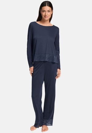 Femme portant un ensemble pyjama bleu marine avec haut à manches longues et pantalon, orné de dentelle sur l’ourlet et les jambes, debout pieds nus sur fond blanc.