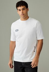 Tricou alb din bumbac cu mâneci scurte, având o aplicație cu un model albastru pe piept și un accent pe cusătura laterală. Purtați cu blugi negri.