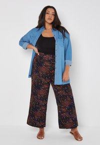 Hellblaues Denim-Hemd über einem schwarzen Tanktop, kombiniert mit weiten Hosen mit einem wirbelnden braunen und marineblauen Muster. Braune Sandalen runden das Outfit ab.