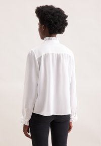 Weiße Bluse mit langen Ärmeln und Rüschkragen, strukturierten vertikalen Streifen, Knopfmanschetten und gerafftem Rückendetail, getragen mit schwarzen Jeans.