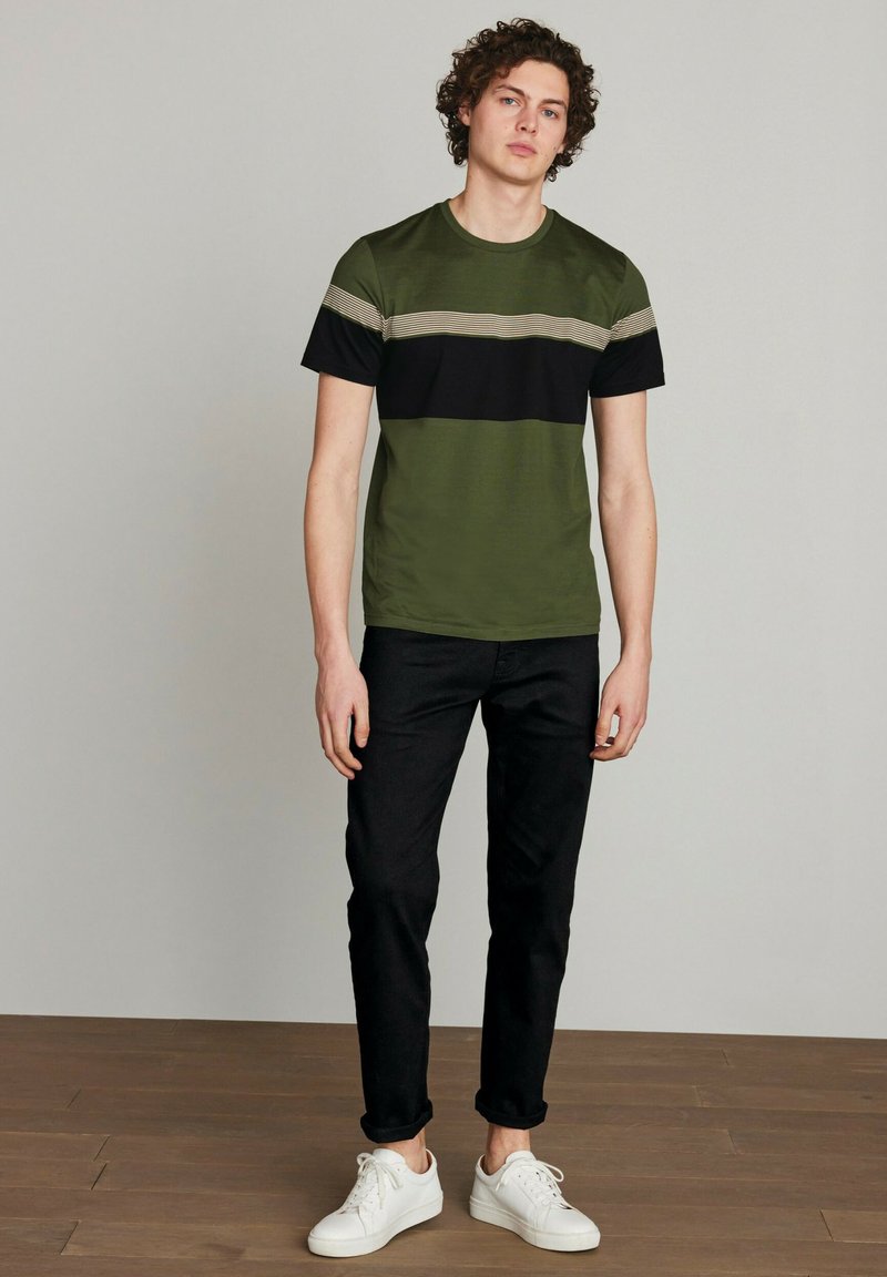 Tricou cu mâneci scurte de culoare verde măsliniu, cu dungi orizontale negre și bej, purtat cu blugi negri și adidași albi. Textură netedă.