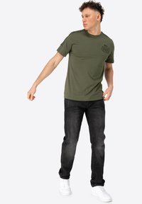 Olivově zelené tričko s kulatým výstřihem a krátkými rukávy, v kombinaci s černými džíny. Bílými teniskami se dotváří outfit, který má detail loga.