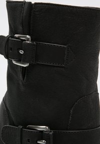 Botín negro de cuero con un acabado texturizado, herrajes de hebilla plateada, diseño de doble correa y forma cónica en la parte superior.