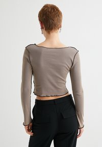 Pitkähiha, cropped-top taupe ribbrakenteisesta kankaasta. Omistaa mustat nyörikuvioiset reunat kaula-aukossa ja hihoissa. Sujuva kangas, tiukka malli.