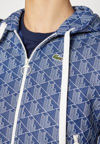 Sweat à capuche zippé à motif bleu avec cordons blancs, porté sur un t-shirt bleu marine, arborant un petit logo crocodile vert sur la poitrine.