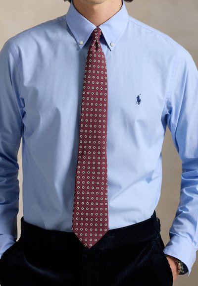 Ανοιχτό μπλε πουκάμισο με κουμπιά και μικρό λογότυπο, συνδυασμένο με μπορντό γραβάτα με γεωμετρικό μοτίβο τετραγώνων. Σκούρο παντελόνι από κορδόνι.