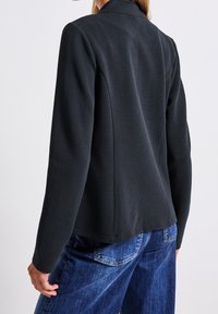 Veste noire texturée avec une coupe structurée, manches longues et col à revers, associée à un jean denim bleu.