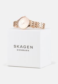 Rozā zelta pulkstenis ar apaļu, minimālistisku seju un saites aproci. Prezentēts baltā kastītē ar melnā krāsā uzdrukātu "SKAGEN DENMARK".