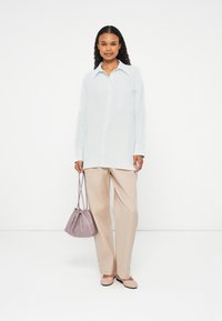 Hellblaues Hemd mit Kragen, kombiniert mit beigen, weit geschnittenen Hosen. Sie hält eine mauvefarbene Zugtasche und trägt hautfarbene Ballerinas.