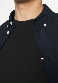 Camisa de botones azul marino sobre una camiseta negra visible en el escote; cuenta con botones blancos y un pequeño logo con acentos en rojo, blanco y azul.