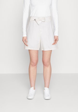 Pantalones cortos beige claro con un detalle de lazo en la cintura, que presentan un ajuste estructurado y dobladillo enrollado. Combinados con zapatillas blancas.