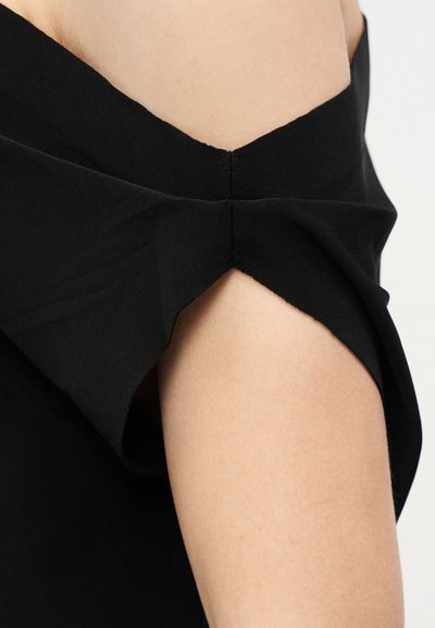 Κοντινό πλάνο μαύρου ενδύματος που αφήνει τους ώμους ελεύθερους, εμφανίζοντας λεπτομέρειες από στριμμένο ύφασμα στο επάνω μέρος του βραχίονα και στον ώμο ενός ατόμου.