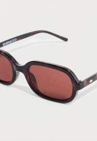 Καφέ γυαλιά ηλίου με χελώνα και βαθμιαία κόκκινα φακούς σε στρογγυλεμένο ορθογώνιο σχήμα. Λεπτοί μαύροι βραχίονες με διακριτική χρυσή λεπτομέρεια.