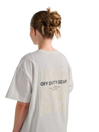 Femme aux cheveux châtain clair coiffés en chignon désordonné, portant un T-shirt ample gris clair avec le texte « OFF DUTY GEAR » dans le dos.