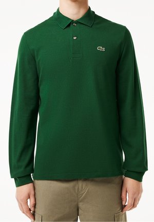 Homme portant un polo à manches longues vert foncé avec deux boutons et un petit logo crocodile sur la poitrine, associé à un pantalon beige.