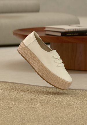 Άσπρο παπούτσι πλατφόρμα με πλέξη από σχοινί τζουτ που αιωρείται πάνω από ένα μπεζ υφαντό χαλί σε μοντέρνο σαλόνι.