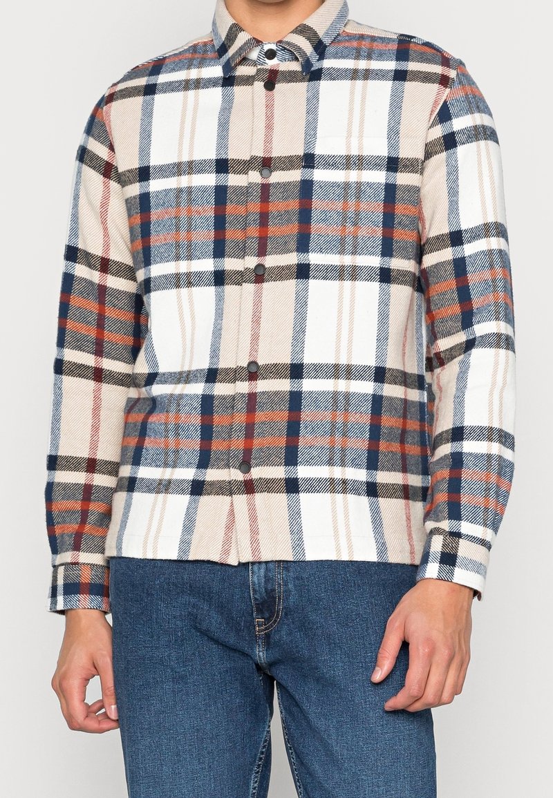 Homme portant une chemise à carreaux boutonnée avec des rayures bleues, blanches, beiges, noires et rouges et un jean en denim bleu.
