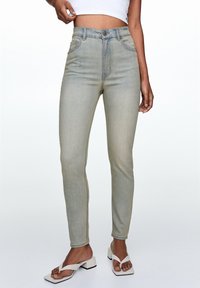 Jeans ajustados de tiro alto en un azul claro con una textura suave, diseño de cinco bolsillos y cierre de botón metálico, combinados con sandalias blancas de tiras.