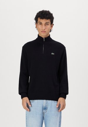 Zwarte pullover met een hoge kraag en ritssluiting, gemaakt van zachte stof, met een klein groen logo op de borst en geribbelde manchetten.