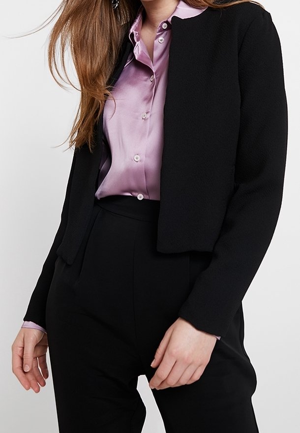 Femme portant un chemisier en satin violet clair rentré dans un pantalon noir taille haute avec un blazer court texturé noir.