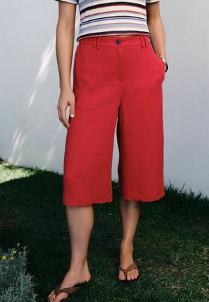 Osoba w czerwonych szerokich, skróconych spodniach, paskowanej bluzce z krótkim rękawem i brązowych klapkach stoi na trawie obok białej ściany.