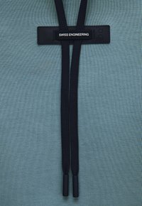 Cordons noirs avec étiquette "SWISS ENGINEERING" sur un fond de tissu bleu sarcelle, vue rapprochée.