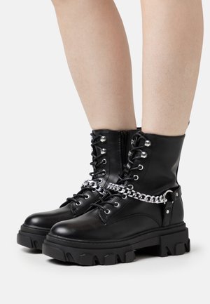 Botas ankle de charol negro con cremallera, parte delantera con cordones, detalle de cadena plateada y suelas gruesas y texturizadas.