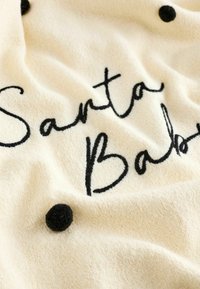 Kremasti pokrivač s crnim izvezenim tekstom koji glasi "Santa Baby" i teksturiranim crnim točkicama po cijelom materijalu.