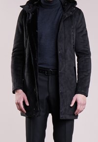 Czarny płaszcz z imitacji zamszu z kapturem, kieszeniami na zamek i miękką, teksturowaną podszewką. Nosi się go na ciemnym golfie i czarnych spodniach.