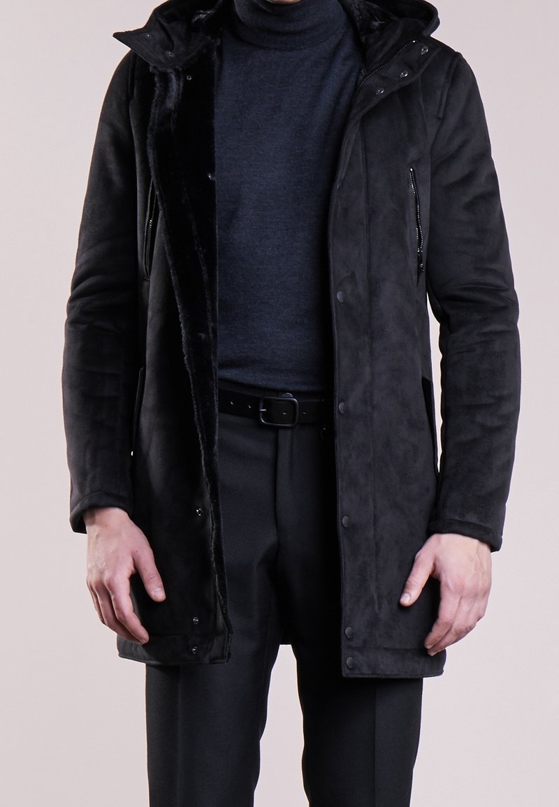 Czarny płaszcz z imitacji zamszu z kapturem, kieszeniami na zamek i miękką, teksturowaną podszewką. Nosi się go na ciemnym golfie i czarnych spodniach.