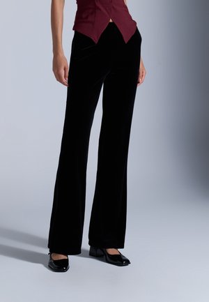 Personne portant un pantalon large en velours noir, des chaussures à talons bas en cuir verni noir et un haut sans manches bordeaux avec un ourlet pointu.