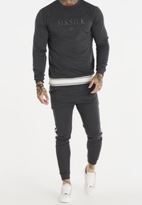 Sudadera gris oscura con logo en relieve. Acompañada de joggers a juego, que presentan rayas laterales y puños elásticos. Llevada con zapatillas blancas.