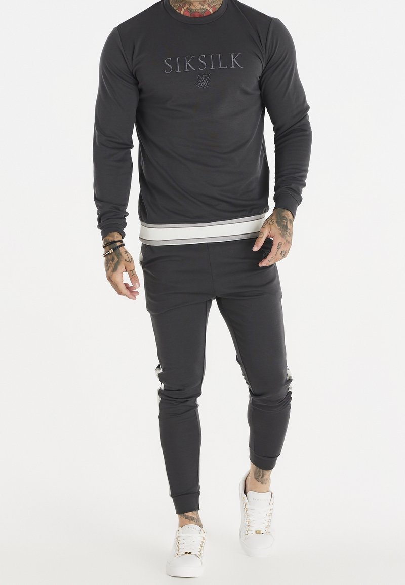Sudadera gris oscura con logo en relieve. Acompañada de joggers a juego, que presentan rayas laterales y puños elásticos. Llevada con zapatillas blancas.
