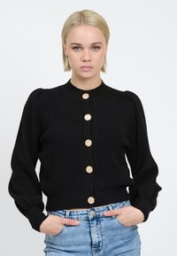 Schwarze Strickjacke mit puffärmel, geripptem Textur und goldenen gestreiften Knöpfen, getragen über hellen Jeansshorts.