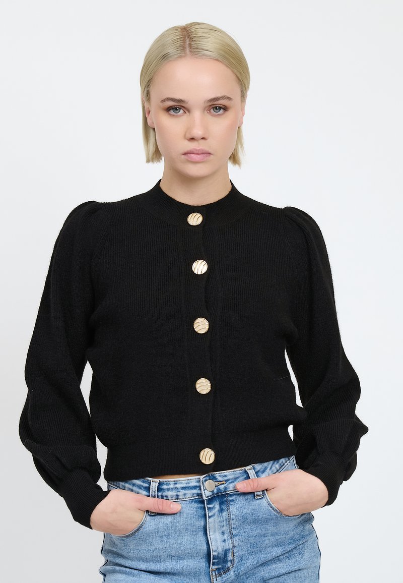 Schwarze Strickjacke mit puffärmel, geripptem Textur und goldenen gestreiften Knöpfen, getragen über hellen Jeansshorts.