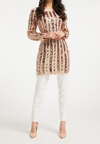 Paillettklänning med vertikala ränder i brons och guld på en mjuk rosa bakgrund, kombinerad med vita byxor och nakna klackar.