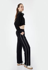 Młoda kobieta z długimi włosami w czarnym krótkim swetrze i czarnych spodniach w białe paski, stojąca z rękami w kieszeniach, w białych trampkach.