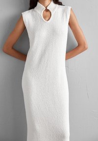 Vestito bianco senza maniche, con texture, colletto alto e scollatura a chiave. Silhouette dritta, lunghezza fino al ginocchio, finitura in tessuto morbido.