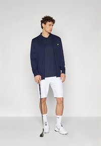 Muškarac u mornarskoj jakni, polo majici, bijelim kratkim hlačama i teniskim cipelama drži palicu za golf, stojeći protiv jednostavne svijetle pozadine.