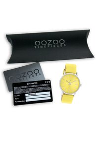 Montre avec un bracelet en silicone jaune, un boîtier rond en argent, un cadran minimaliste et une carte de garantie noire à côté d'une boîte de marque.