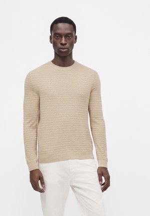 Homme portant un pull beige texturé à manches longues et un pantalon blanc, debout devant un fond blanc uni.