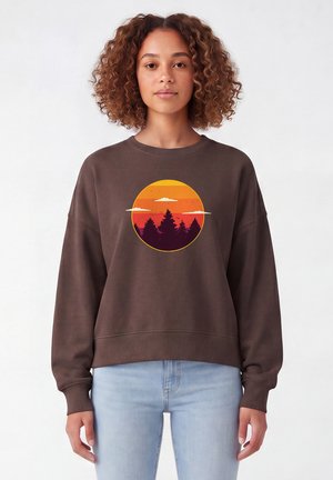 Frau mit lockigem Haar, die einen braunen Sweatshirt mit Sonnenuntergangs- und Baumsilhouette-Design trägt, kombiniert mit hellblauen Jeans, steht vor weißem Hintergrund.