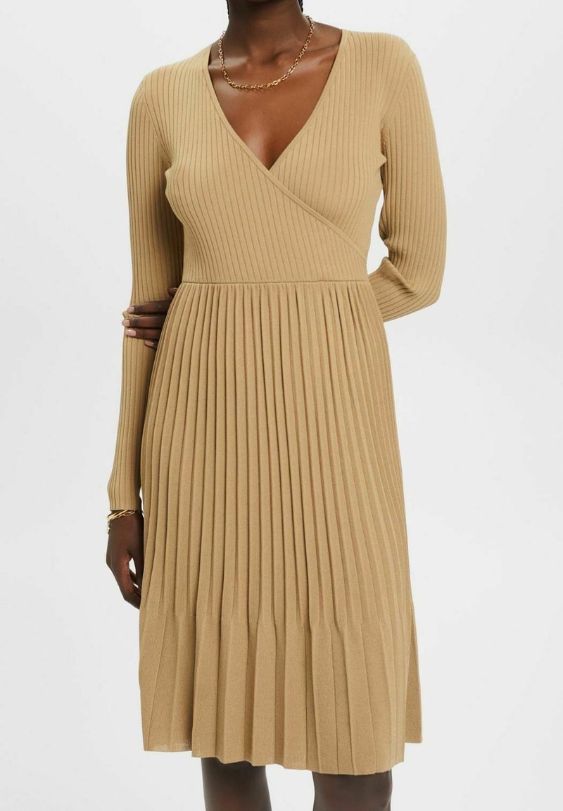 Knä-lång, ribbad stickad klänning i tan med långa ärmar, djup V-halsringning och samlade plisseringar som börjar vid midjan för textur och rörelse.