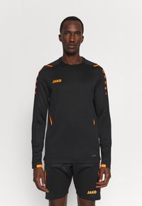 Maglia sportiva nera a maniche lunghe con accenti arancioni e logo "JAKO". Presenta un design elegante e un materiale testurizzato. Abbinata a pantaloni corti neri.