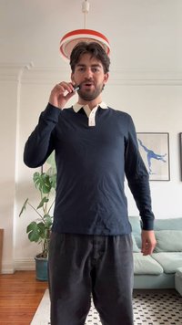 Jūros laivyno ilgas rankovėmis marškiniai su balta apykakle, juodos kelnės, laikantis vapingą. Patalpos aplinka su žaliu augalu ir šviesios spalvos sofa.