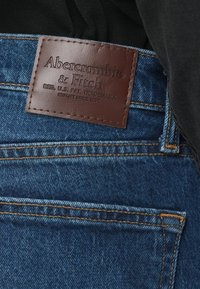 Dongeribukser i en mørkeblå farge, med en brun skinnlapp preget med "Abercrombie & Fitch" og kontrasterende oransjefargede sømmer.