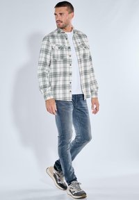 Karierte Flanellhemd in Weiß und Grün, zwei Vordertaschen, Knopfdesign, über einem weißen T-Shirt getragen, kombiniert mit blauen Jeans und Turnschuhen.