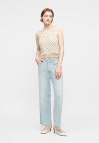 Γυναίκα που φοράει μπεζ τοπ χωρίς μανίκια με δίχτυ, ανοιχτό μπλε ψηλόμεσο τζιν και λευκά παπούτσια με τακούνι, στέκεται με το ένα χέρι στην τσέπη.