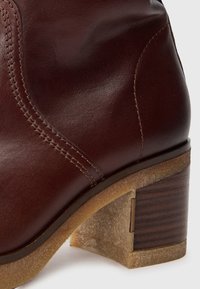 Bottine en cuir marron avec une finition lisse, des détails de couture verticaux et un talon épais en texture bois de couleur beige.
